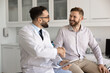 © fizkes - Happy male doctor and patient shaking hands in hospital examination room, sitting on medical couch, giving handshake, smiling, laughing. Man thanking physician for help, support