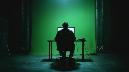 Person sitting at their desk, facing away from the camera with an illuminated green screen in front of them