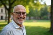© Viktor - Portrait of smiling 65-year-old man with glasses in park. He has grey hair and beard, wearing casual striped shirt. Background shows blurred green lawn and trees, suggesting relaxed outdoor setting.