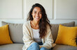 © Viktor - Cheerful young Latin American woman smiles warmly from a cozy home sofa. Dressed casually in jeans and a knit cardigan, she radiates happiness and positive energy while relaxing indoors.