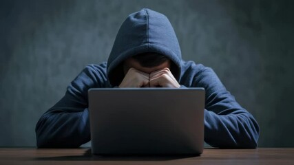 Person hoodie sitting at table with laptop indoors, covering face with hands showing frustration and stress, technology and computer use in casual setting, tired and isolated at work desk