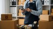 © Muhammad - Person in apron using phone, surrounded by shipping boxes, preparing orders.