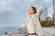 © SHOTPRIME STUDIO - woman enjoying calm moment outdoors near sea with eyes closed, wearing light jacket and white blouse, urban background, peaceful expression, natural light, emotional relaxation