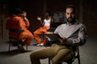 © pressmaster - Caucasian middle aged man holding clipboard sitting in foreground, leading group therapy session with diverse young adults and teenagers in prison uniforms talking in background
