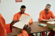 © pressmaster - Black man holding handwritten letter standing in foreground while two men in orange uniforms sitting at table in background listening attentively during group activity