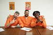 © pressmaster - Three young adult inmates sitting at table wearing orange uniforms, smiling and posing together, one making peace sign, paperwork and pens on table