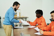 © pressmaster - Middle aged Caucasian man handing documents to two male prisoners in orange uniforms participating in educational activity in prison classroom setting