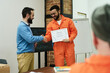 © pressmaster - Middle aged man congratulating young adult man in orange prison uniform receiving certificate during educational program in classroom setting with another inmate observing