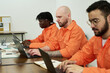 © pressmaster - Three young adult men sitting at desks using laptops and writing in notebooks, participating in educational program inside prison classroom