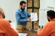 © pressmaster - Middle aged Caucasian man instructing two adult men in orange uniforms during classroom session, holding printed worksheet while explaining educational material in prison setting