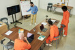 © pressmaster - Multiethnic group of young adult men wearing orange prison uniforms sitting at tables using laptops, while male instructor explaining lesson on whiteboard in classroom setting