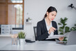 © MINAE - Young Asian businesswoman sitting at a modern desk, holding a pen and analyzing financial paperwork while working on a laptop in a contemporary office environment