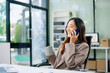 © laddawan - Confident young Asian businesswoman smiles while multitasking talking on her smartphone and using her laptop at a modern office desk