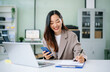 © laddawan - Smiling young Asian businesswoman multitasks at her office desk working on laptop while checking smartphone