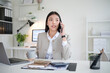 © wattana - Young businesswoman talking on the phone while holding a coffee cup at desk in a modern office.