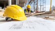 © BANDASAK - A yellow hard hat on blueprints at a construction site with workers in the background focusing on their tasks.