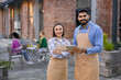 © sofiko14 - A diverse pair of restaurant staff members smile at the camera outside, while customers dine in the background.