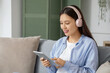 © Pixel-Shot - Beautiful young Asian woman in headphones listening to audiobook with modern tablet computer and sitting on sofa in living room