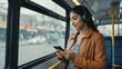 © VortexX - Smiling woman with headphones using smartphone on bus wearing tan jacket