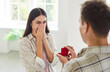 © Studio Romantic - Young man with a red gift box proposing to his happy girlfriend standing in living room at home. Male person giving surprise gift with a engagement ring or jewelry to his happy pretty girlfriend.