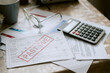 © AnnaStills - Senior Caucasian couple struggling with overdue utility bills, holding documents marked past due on cluttered table with calculator and eyeglasses, financial hardship evident