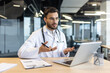 © Liubomir - A doctor in a white coat uses a laptop for a video call, consulting or telemedecine, in a modern office setting.