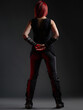 © Loginova - A rear view of a woman with crimson hair wearing a black outfit in a dimly lit environment. Stylish details like buckled boots emphasize a unique and bold fashion sense.