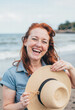© Eliane - A joyful redhead woman with a genuine, exuberant laugh holding a straw hat at the beach, capturing a carefree and happy moment on a sunny summer vacation.