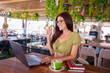 © Javier - Young woman waving at her laptop while making a video call and drinking a cappuccino in a restaurant