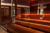 Modern wooden sauna with warm lighting behind a glass wall, located near an indoor pool with water reflections and relaxing spa atmosphere