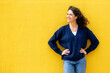 © mimagephotos - Confident woman smiling with hands on hips, standing against a bold yellow wall
