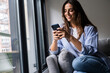 © simona - Young woman casually using her smartphone at home. A modern lifestyle filled with chatting, browsing, booking, online shopping and managing daily tasks in the digital world of a connected generation
