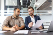 © Liubomir - Two businessmen reviewing documents at a table in a modern office setting. The man on the right is pointing at the paper with a pen.