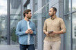 © Liubomir - Two businessmen conversing outdoors near a modern glass building, one holding a coffee cup, the other a tablet.