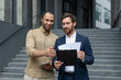 © Liubomir - Two businessmen review documents outside a modern building, discussing business strategy and collaboration.