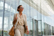 © Alina - Businesswoman in beige suit walking confidently outside modern glass building during daytime