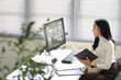 © Prathankarnpap - Asian woman participating in an online team meeting while reviewing documents at her desk