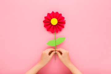 Wall Mural - Little child girl hands holding and showing red flower with green leaves from colorful application paper on light pink table background. Pastel color. Closeup. Point of view shot. Top down view.