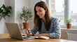 © MstShilpi12 - Young woman smiling while working on a laptop and writing notes in a notebook at a wooden desk