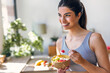 © nenetus - Smiling sporty woman eating a healthy fruit bowl while sitting in the kitchen at home