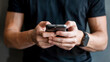© Shamim - Man using smartphone for texting and browsing, wearing a black smartwatch, close-up shot showing the hands and device in a lifestyle tech gadget scene against a gray wall.