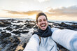 © Davide Angelini - Handsome young man taking selfie pic with smart mobile phone outdoors - Traveler guy smiling at camera with sunset on background - Traveling life style and technology concept
