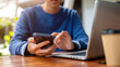 © bongkarn - Close up of man hands holding and typing on phone aside laptop at wooden table in cafe or canteen.
