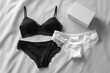 © Nyxoria - Elegant lingerie set with a black lace-trimmed bra and panties, accompanied by a white pair, presented on a silky surface next to a gift box.