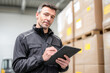 © Kowit - A man in a black jacket writes on a clipboard while inspecting stacked cardboard boxes in a warehouse setting.