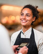 © Kowit - Smiling female barista in an apron interacts with a customer using a card payment device in a warmly lit cafe setting.