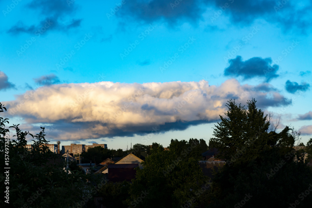 Panoramic view of residential houses and trees under large illuminated cumulus cloud in blue sky during sunset. Urban landscape and weather observation concept. Design for poster, wallpaper, banner. W
