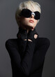 © SHOTPRIME STUDIO - Stylish young woman with short blonde hair wearing oversized sunglasses and black gloves, striking a confident pose against a dark grey background.