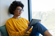 © Wavebreak Media - African American woman sitting by large window wearing yellow knit sweater using tablet, copy space