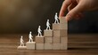 © Sina Blaze - Conceptual image of human development, leadership, and career growth showing paper cutout figures ascending wooden blocks as a hand places the final step, symbolizing success and support.